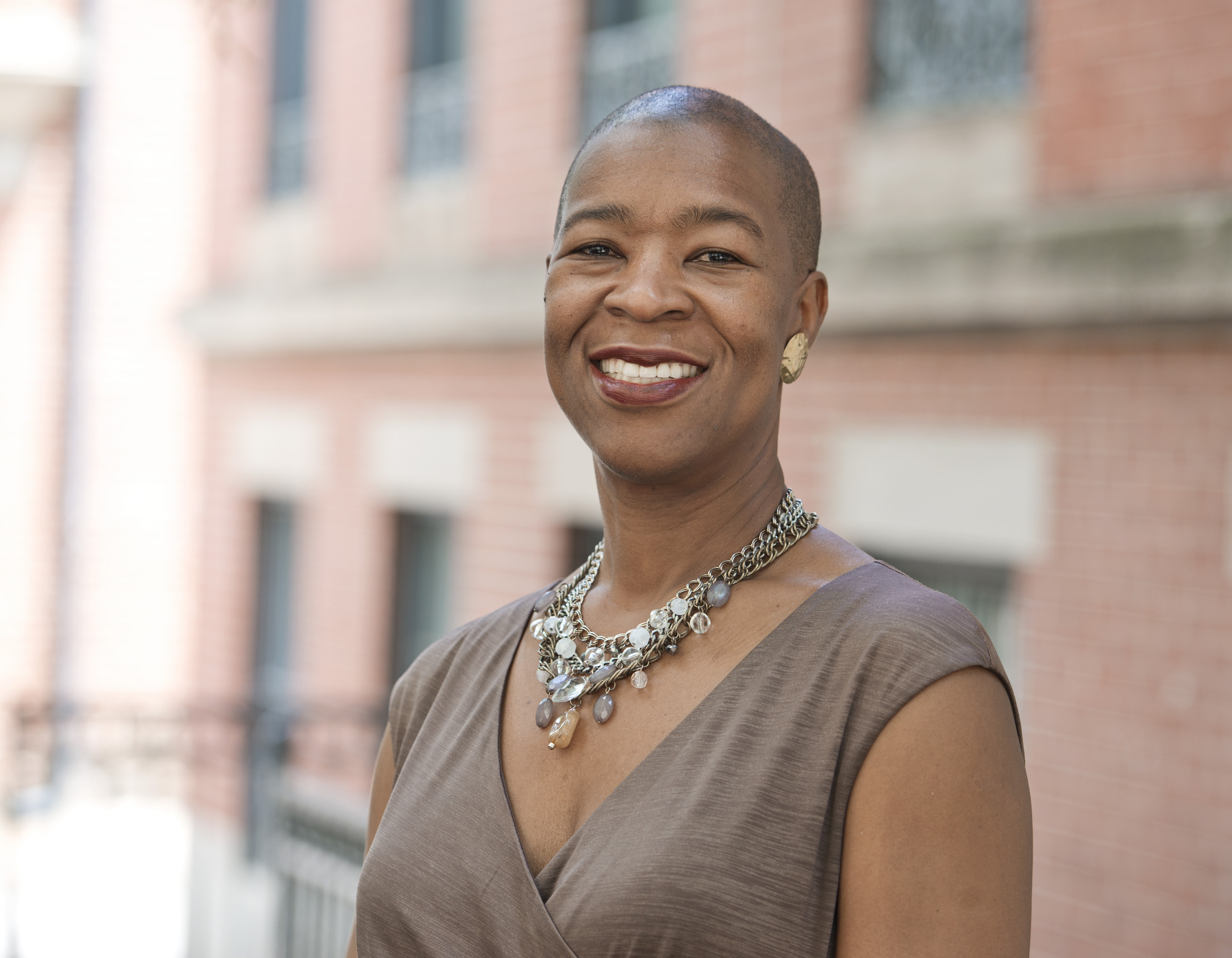 Headshot of Danista E. Hunte: Danista is a smiling Black woman of Caribbean descent with a bald head, wearing a short sleeved brown top, a metal and beaded statement necklace, and large round gold earrings. She is standing in front of a brick building.