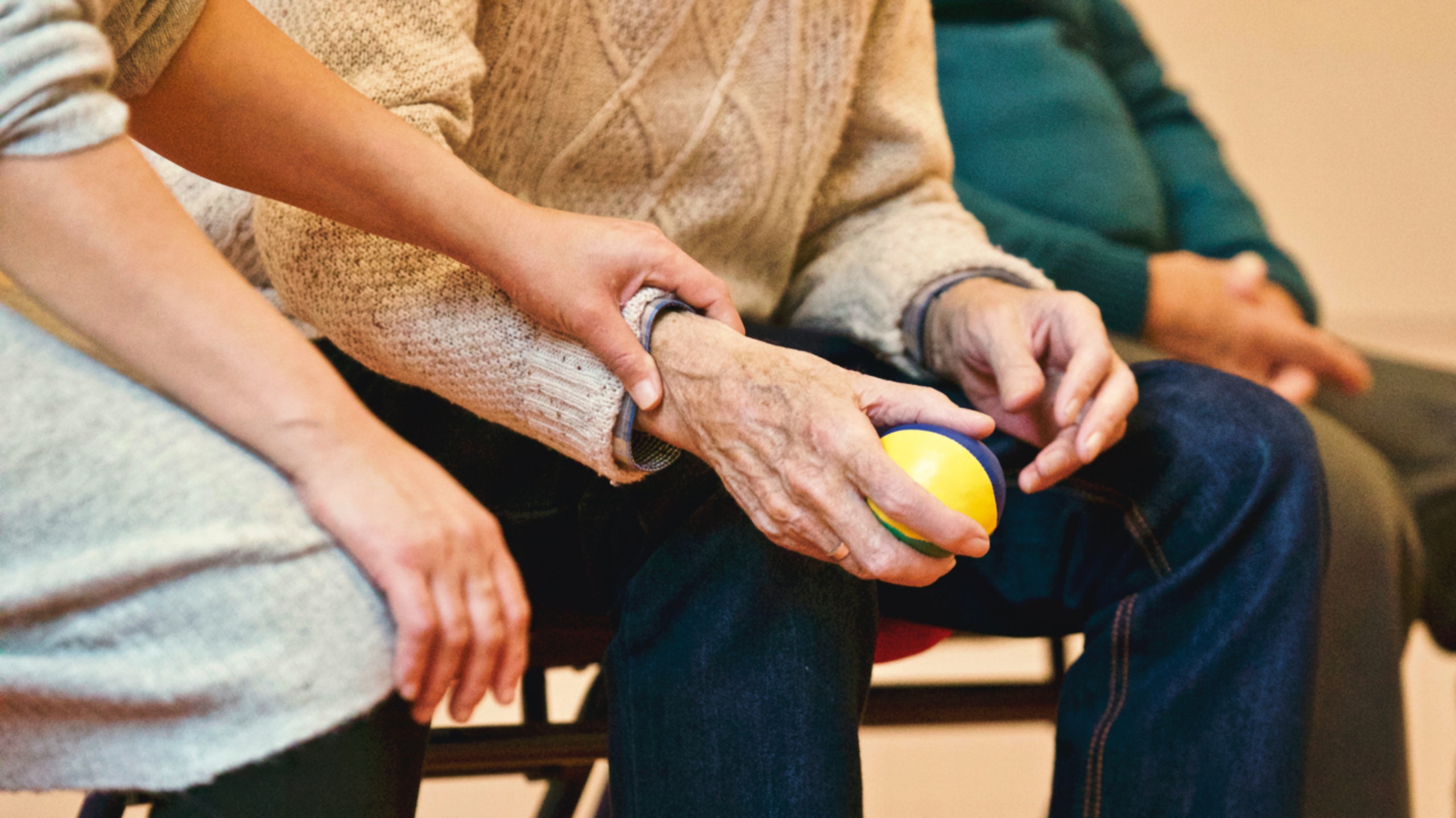 older adult holding a ball another non older adult is holding their wrist for assitance